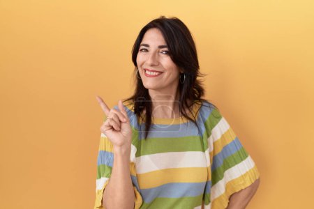 Photo for Middle age hispanic woman standing over yellow background with a big smile on face, pointing with hand and finger to the side looking at the camera. - Royalty Free Image