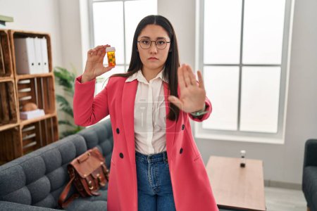 Photo for Young latin woman working as therapist holding jar of pills with open hand doing stop sign with serious and confident expression, defense gesture - Royalty Free Image