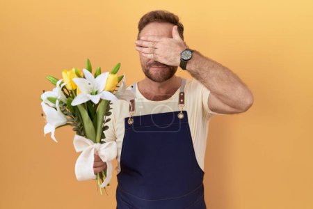Téléchargez les photos : Homme du Moyen Age avec magasin de fleuriste barbe tenant des fleurs souriant et riant avec la main sur le visage couvrant les yeux pour la surprise. concept aveugle. - en image libre de droit
