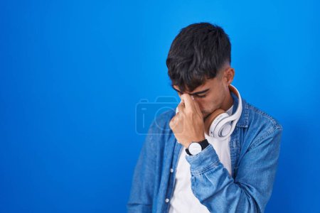 Foto de Joven hispano de pie sobre fondo azul cansado frotando nariz y ojos sintiendo fatiga y dolor de cabeza. concepto de estrés y frustración. - Imagen libre de derechos