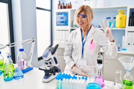 Téléchargez les photos : Femme blonde d'âge moyen travaillant au laboratoire scientifique montrant et pointant vers le haut avec les doigts numéro quatre tout en souriant confiant et heureux. - en image libre de droit