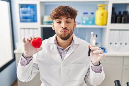 Photo for Arab man with beard working at scientist laboratory holding blood samples looking at the camera blowing a kiss being lovely and sexy. love expression. - Royalty Free Image