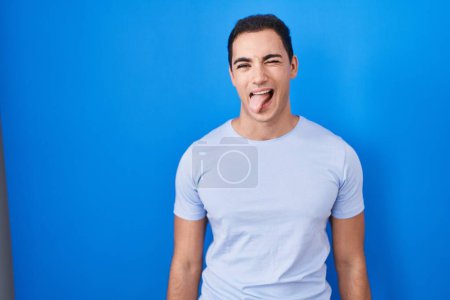 Téléchargez les photos : Jeune homme hispanique debout sur fond bleu langue collante heureux avec une expression drôle. concept d'émotion. - en image libre de droit