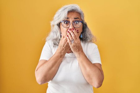 Foto de Middle age woman with grey hair standing over yellow background shocked covering mouth with hands for mistake. secret concept. - Imagen libre de derechos