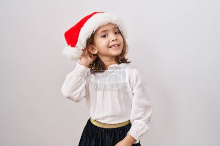 Photo for Little hispanic girl wearing christmas hat smiling with hand over ear listening and hearing to rumor or gossip. deafness concept. - Royalty Free Image