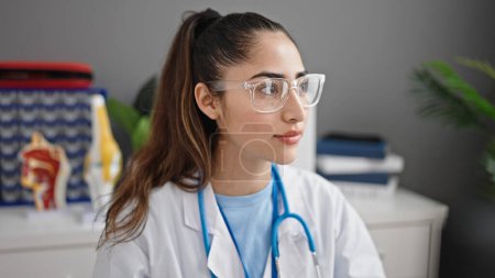 Photo for Young beautiful hispanic woman doctor sitting on table looking to the side at clinic - Royalty Free Image