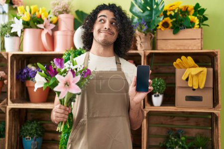 Foto de Hombre hispano con el pelo rizado trabajando en floristería mostrando la pantalla del teléfono inteligente sonriendo mirando hacia un lado y mirando hacia otro pensando. - Imagen libre de derechos