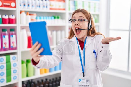 Photo for Redhead woman working at pharmacy drugstore doing video call with tablet celebrating achievement with happy smile and winner expression with raised hand - Royalty Free Image