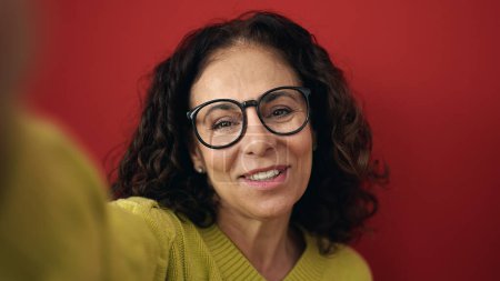 Téléchargez les photos : Moyen Age hispanique femme souriante confiante faire selfie par caméra sur fond rouge isolé - en image libre de droit