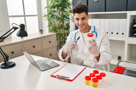 Photo for Young hispanic man wearing doctor uniform pointing to analysis test tube at clinic - Royalty Free Image