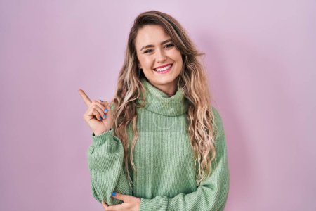 Téléchargez les photos : Jeune femme caucasienne debout sur fond rose avec un grand sourire sur le visage, pointant avec la main et le doigt vers le côté regardant la caméra. - en image libre de droit