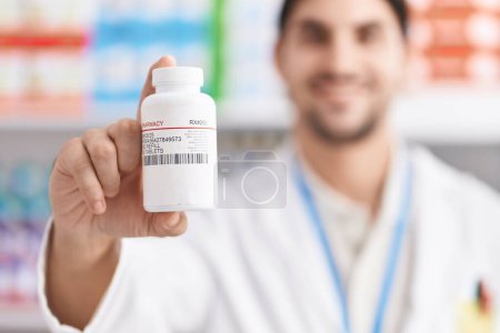 Téléchargez les photos : Young hispanic man pharmacist smiling confident holding pills bottle at pharmacy - en image libre de droit
