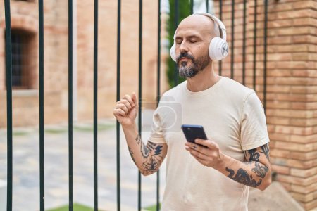 Photo for Young bald man listening to music and dancing with serious expression at street - Royalty Free Image