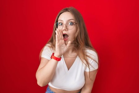 Téléchargez les photos : Jeune femme caucasienne debout sur fond rouge main sur bouche racontant rumeur secrète, chuchotant conversation malicieuse - en image libre de droit