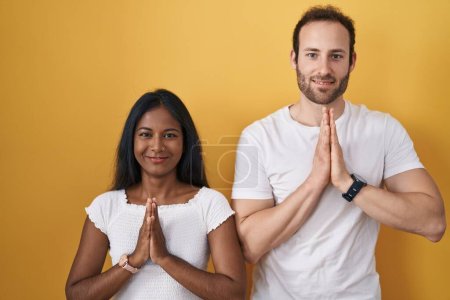 Photo for Interracial couple standing over yellow background praying with hands together asking for forgiveness smiling confident. - Royalty Free Image