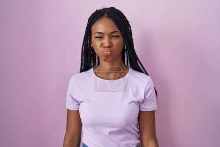 Foto de Mujer afroamericana con trenzas de pie sobre fondo rosa hinchando mejillas con cara divertida. boca hinchada de aire, expresión loca. - Imagen libre de derechos