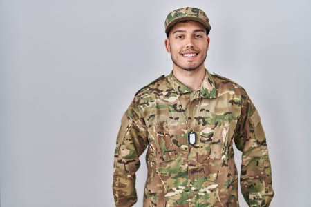 Foto de Joven hombre hispano vistiendo uniforme camuflaje del ejército con una sonrisa alegre y fresca en la cara. persona afortunada. - Imagen libre de derechos