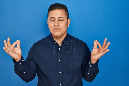 Téléchargez les photos : Jeune homme hispanique debout sur fond bleu se détendre et sourire les yeux fermés en faisant geste de méditation avec les doigts. concept de yoga. - en image libre de droit