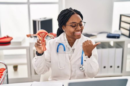 Foto de Mujer médica afroamericana sosteniendo modelo anatómico de órgano genital femenino apuntando el pulgar hacia el lado sonriendo feliz con la boca abierta - Imagen libre de derechos