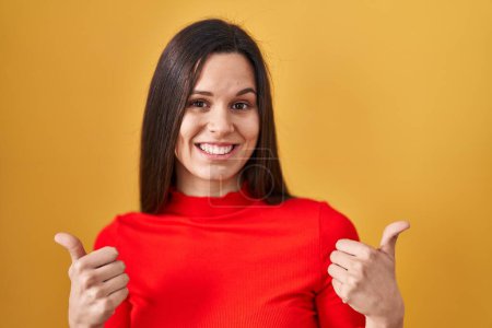 Photo for Young hispanic woman standing over yellow background success sign doing positive gesture with hand, thumbs up smiling and happy. cheerful expression and winner gesture. - Royalty Free Image