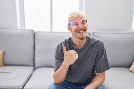 Téléchargez les photos : Jeune homme caucasien portant des lunettes de réalité virtuelle souriant heureux et positif, pouce levé faire excellent et signe d'approbation - en image libre de droit