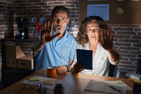 Photo for Middle age hispanic couple using touchpad sitting on the table at night doing stop gesture with hands palms, angry and frustration expression - Royalty Free Image