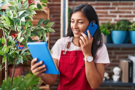 Téléchargez les photos : Jeune belle hispanique femme fleuriste parler sur smartphone en utilisant touchpad à la boutique de fleurs - en image libre de droit