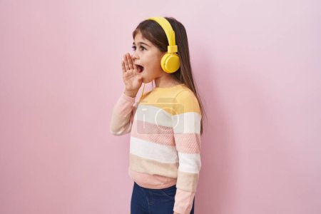 Téléchargez les photos : Petite fille hispanique écoutant de la musique à l'aide d'écouteurs désemparés et confus avec les bras ouverts, aucune idée et visage douteux. - en image libre de droit