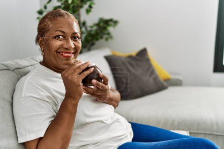 Foto de Senior africana americana mujer sonriendo confiado beber mate infusión en casa - Imagen libre de derechos