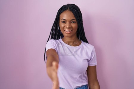 Foto de Mujer afroamericana con trenzas de pie sobre fondo rosa sonriendo amistoso ofreciendo apretón de manos como saludo y bienvenida. negocios exitosos. - Imagen libre de derechos