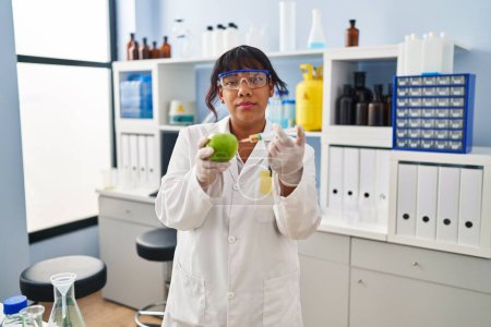 Photo for Hispanic woman working at scientist laboratory with apple relaxed with serious expression on face. simple and natural looking at the camera. - Royalty Free Image