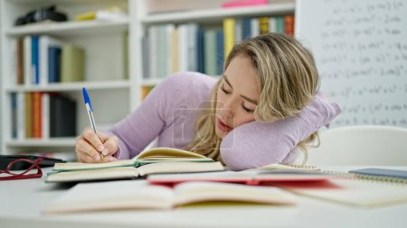Téléchargez les photos : Jeune femme blonde étudiante fatiguée à écrire des notes en classe - en image libre de droit