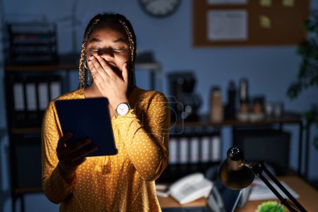 Téléchargez les photos : Femme afro-américaine avec des tresses travaillant au bureau la nuit avec une tablette ennuyeuse bâillant fatigué couvrant la bouche avec la main. agitation et somnolence. - en image libre de droit