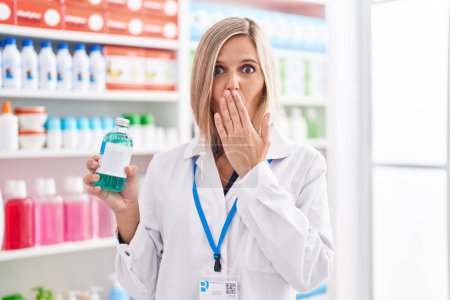 Téléchargez les photos : Femme blonde travaillant à la pharmacie droguerie tenant du sirop couvrant la bouche avec la main, choquée et effrayée par l'erreur. expression surprise - en image libre de droit