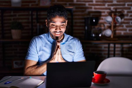 Téléchargez les photos : Homme hispanique avec barbe à l'aide d'un ordinateur portable la nuit priant avec les mains ensemble demandant pardon souriant confiant. - en image libre de droit