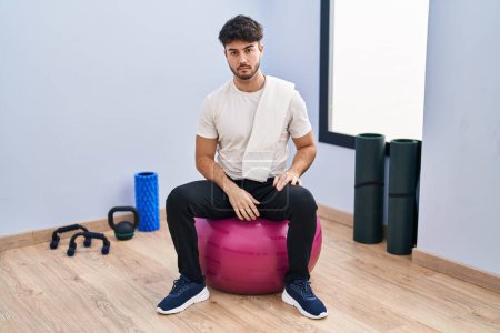 Photo for Hispanic man with beard sitting on pilate balls at yoga room relaxed with serious expression on face. simple and natural looking at the camera. - Royalty Free Image