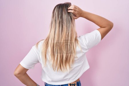 Téléchargez les photos : Jeune femme blonde debout sur fond rose vers l'arrière pensant au doute avec la main sur la tête - en image libre de droit