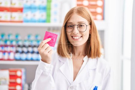 Téléchargez les photos : Jeune rousse femme travaillant à la pharmacie pharmacie tenant préservatif regardant positif et heureux debout et souriant avec un sourire confiant montrant des dents - en image libre de droit