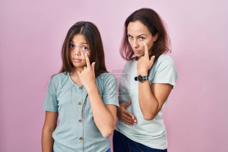 Foto de Joven madre e hija de pie sobre fondo rosa apuntando al ojo observándote gesto, expresión sospechosa - Imagen libre de derechos