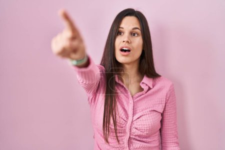 Foto de Young hispanic woman standing over pink background pointing with finger surprised ahead, open mouth amazed expression, something on the front - Imagen libre de derechos