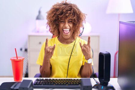 Photo for Young hispanic woman with curly hair playing video games wearing headphones shouting with crazy expression doing rock symbol with hands up. music star. heavy concept. - Royalty Free Image