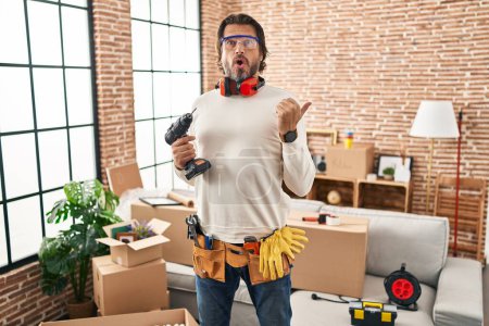 Photo for Handsome middle age man holding screwdriver at new home surprised pointing with hand finger to the side, open mouth amazed expression. - Royalty Free Image