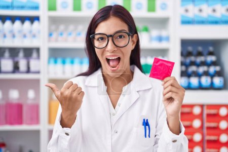 Téléchargez les photos : Jeune femme hispanique travaillant à la pharmacie tenant préservatif pointant le pouce vers le côté souriant heureux avec la bouche ouverte - en image libre de droit