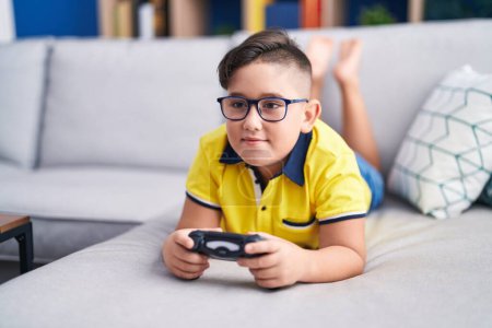 Foto de Joven niño hispano jugando videojuegos sosteniendo el controlador en el sofá sonriendo con una sonrisa alegre y fresca en la cara. mostrando dientes. - Imagen libre de derechos