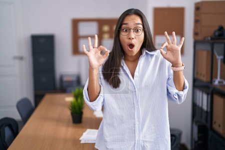 Téléchargez les photos : Jeune femme hispanique au bureau à l'air surpris et choqué de faire ok symbole d'approbation avec les doigts. expression folle - en image libre de droit