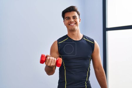 Foto de Joven hombre hispano sonriendo confiado usando entrenamiento de mancuerna en el centro deportivo - Imagen libre de derechos