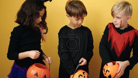 Photo for Group of kids wearing halloween costume putting sweets in pumpkin basket over isolated yellow background - Royalty Free Image