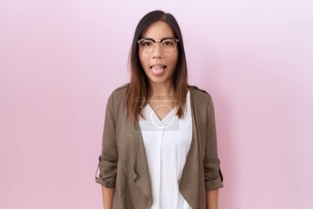 Téléchargez les photos : Femme chinoise du Moyen Âge portant des lunettes sur fond rose langue collante heureux avec une expression drôle. concept d'émotion. - en image libre de droit