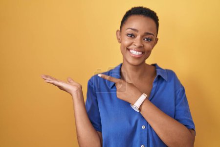 Téléchargez les photos : Femme afro-américaine debout sur fond jaune étonné et souriant à la caméra tout en présentant avec la main et pointant du doigt. - en image libre de droit