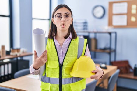 Photo for Hispanic girl architect holding build project blueprints making fish face with mouth and squinting eyes, crazy and comical. - Royalty Free Image